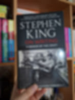 Hand holding "On Writing" by Stephen King. Cover shows a black-and-white image of a person writing with a dog nearby. Bookshelf in background.