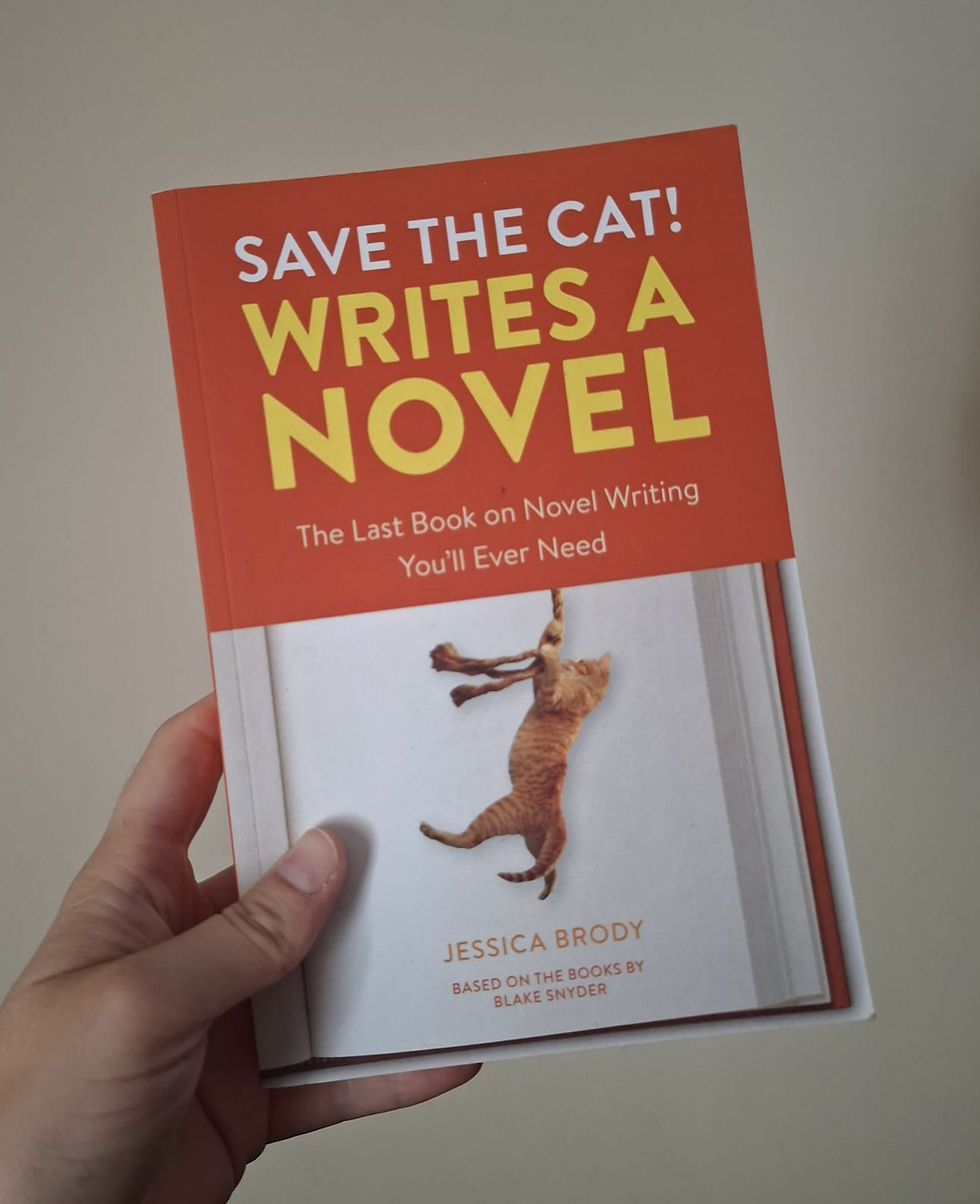 Hand holding the book "Save the Cat! Writes a Novel" by Jessica Brody. Cover shows a cat hanging, on a red-orange background.