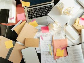 Laptop on cluttered desk with open books, notebooks, and scattered sticky notes. Papers show math equations. Bright, busy atmosphere.