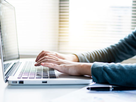 Hands typing on a silver laptop at a bright desk, with blinds in the background and a pen in the foreground.