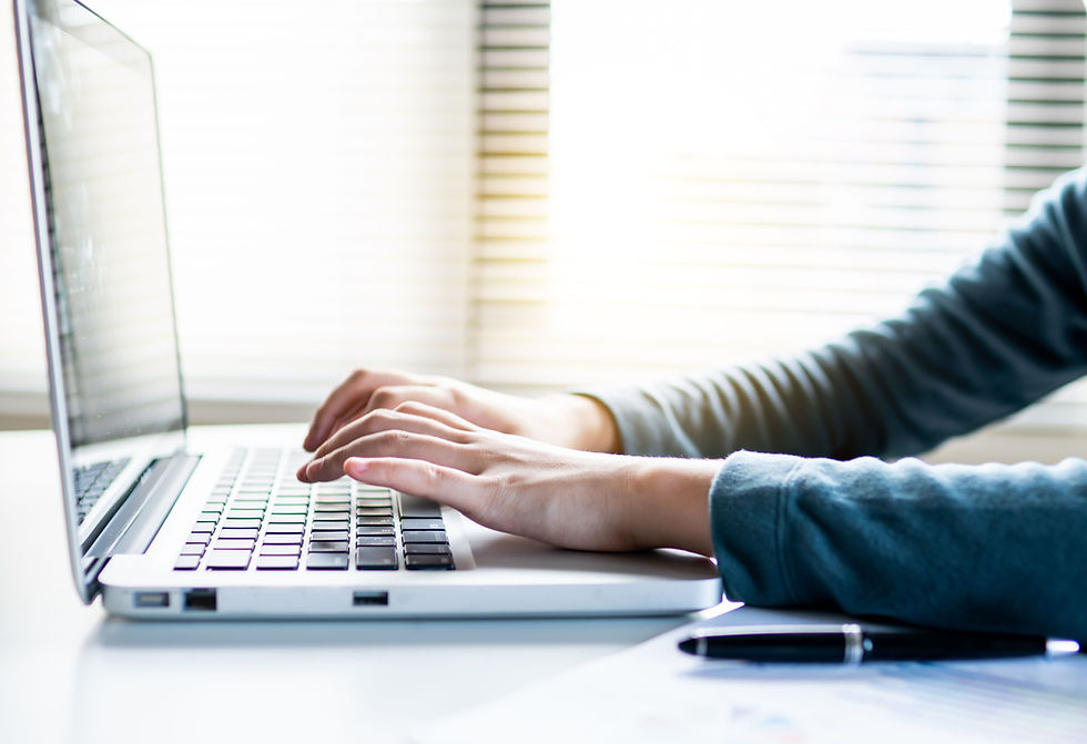 Hands typing on a silver laptop at a bright desk, with blinds in the background and a pen in the foreground.