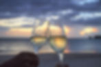 Two champagne glasses clink against a beach sunset backdrop, creating a celebratory mood with the ocean and colorful sky in view.
