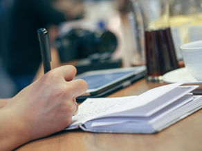 Hand writing in a notebook at a wooden table with a pen. Nearby, a tablet, coffee cup, and drinks create a thoughtful atmosphere.
