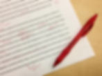 Printed essay with red pen edits, including underlines and notes. A red pen lies on a wooden desk beside the paper.