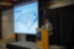 North Carolina Department of Transportation representative presents at podium with laptop, showing North Carolina Rail System map on a projector screen. Black curtains and chairs in background.