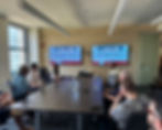 People seated in a conference room watching screens displaying "Race to Chicago Roundtable." Office setting with large windows.