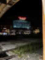 Night view of Lucas Oil Stadium, lit up with bright red and white signage. Dimly lit foreground features railway tracks and gravel.