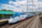 Blue and white high-speed Acela train at a station under a cloudy sky, with a green-roofed platform and tracks stretching into the distance.