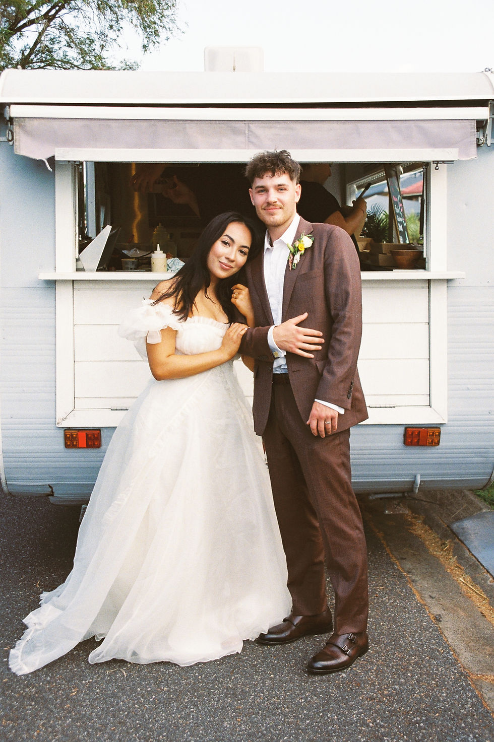 Bride in a white gown and groom in a brown suit stand smiling in front of a white food truck, with trees in the background.