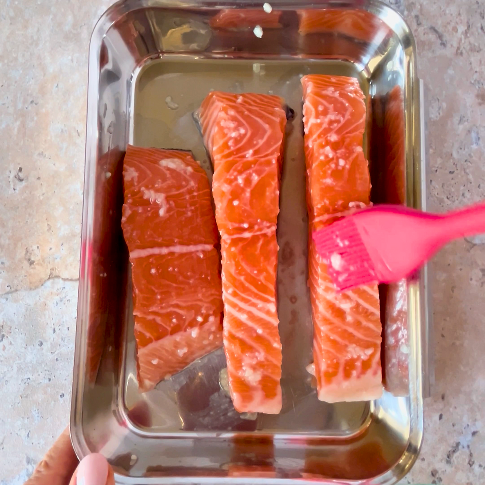 Three salmon fillets in a metal tray being brushed with salt koji using a pink brush. The background is a textured, light-colored surface.