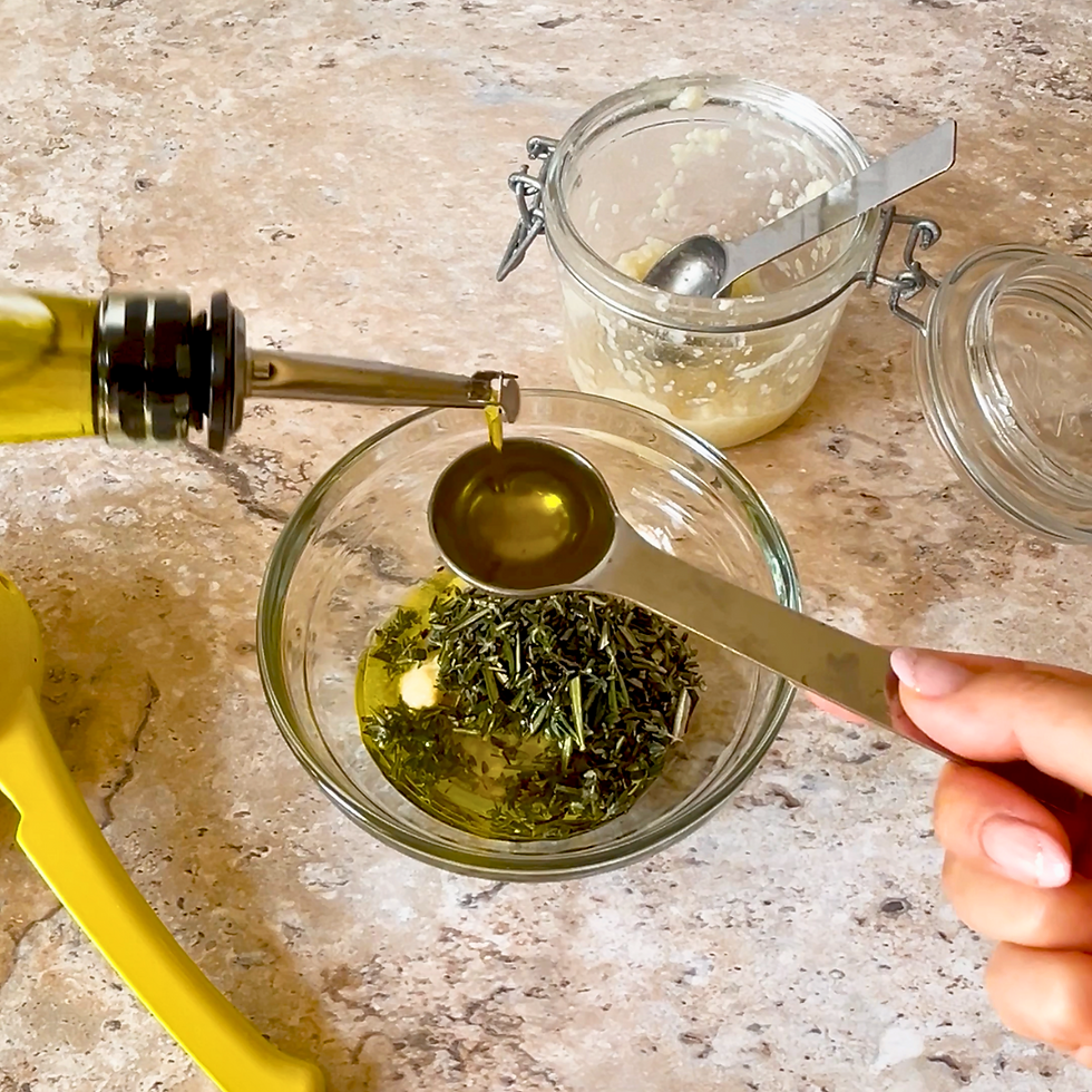 Olive oil is poured into a measuring spoon over a bowl with herbs. A jar of salt koji paste is in the background on a marble surface.