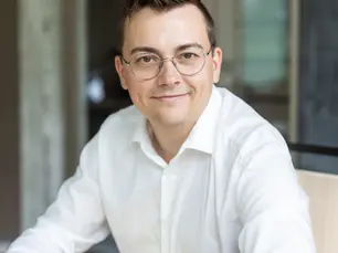 Felix Fröhlich, Versicherungsmakler aus Potsdam, sitzt lächelnd am Schreibtisch mit Laptop und Brille im weißen Hemd.