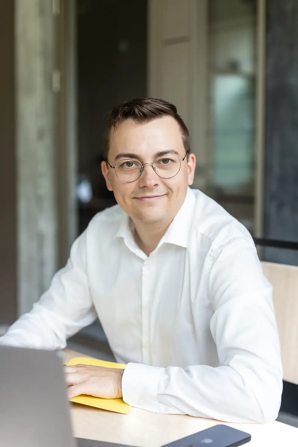 Felix Fröhlich, Versicherungsmakler aus Potsdam, sitzt lächelnd am Schreibtisch mit Laptop und Brille im weißen Hemd.