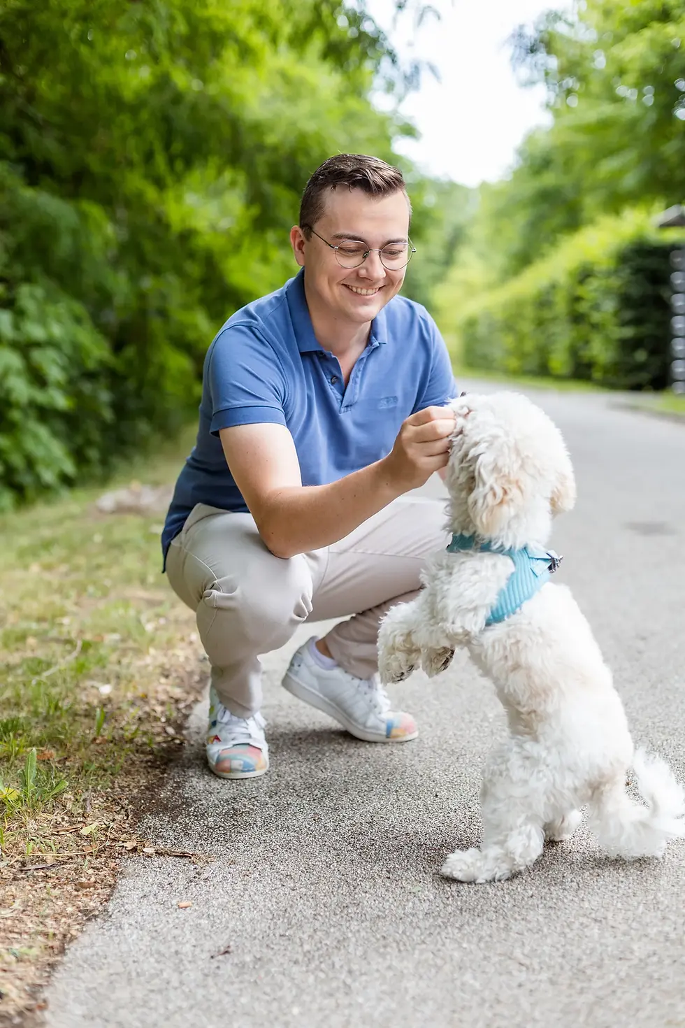 Felix Fröhlich hockt lächelnd auf einem Weg und streichelt seinen weißen Hund Teddy mit türkisem Geschirr, im Hintergrund grüne Bäume