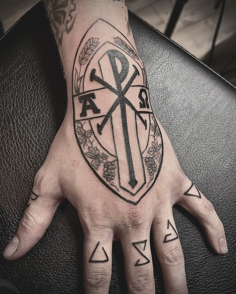 Geometrisches Blackwork-Tattoo auf dem Handrücken mit dem christlichen Chi-Rho-Symbol (Chrismon) sowie Alpha und Omega, kombiniert mit Runen-Symbolen auf den Fingern, gestochen bei Neuanstich in Horneburg Landkreis Stade.