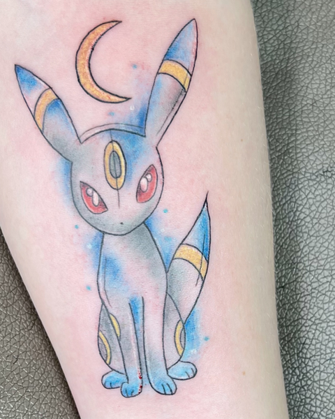 Buntes Anime-Tattoo des Unlicht-Pokémons Nachtara (Umbreon) mit blauem Aquarell-Hintergrund und Mondsichel, gestochen bei Neuanstich in Horneburg nahe Stade.