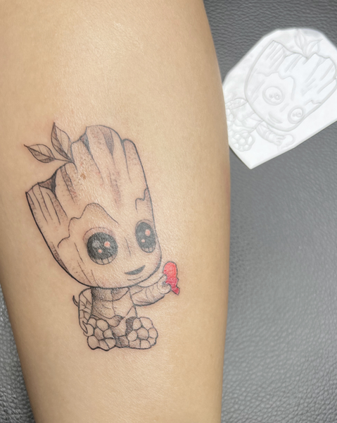 Kleines, detailreiches Baby Groot Tattoo im Comic-Stil auf dem Arm, der ein kleines rotes Herz in der Hand hält, gestochen bei Neuanstich in Horneburg Landkreis Stade