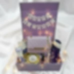Luxury purple birthday gift box for her with a "Happy Birthday" banner. Includes a quilted lavender crossbody purse with a gold chain, a "The Curated Chest" jar chocolate, premium skincare, and fairy lights. High-end personalized hamper idea for women.