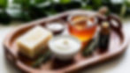 vibrant display of ingredients like Castile soap, coconut milk, honey, and rosemary essential oil on a wooden tray, with greenery in the background to emphasize a natural and nourishing theme.