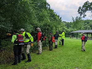 Search and rescue team members in high-visibility gear training outdoors, lined up along a wooded edge while conducting a coordinated search.