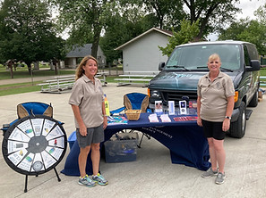 Public Preparedness Group volunteers at a community event, standing by an outreach table with preparedness materials, a prize wheel, and a van in the background.