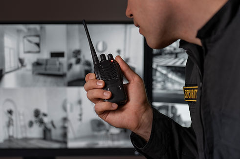 A security guard is talking into a walkie-talkie. In the background, a monitor displays multiple security camera feeds.