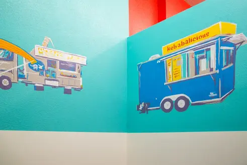 Two food trucks, one says 'Kebabalicious' on a turquoise wall background.