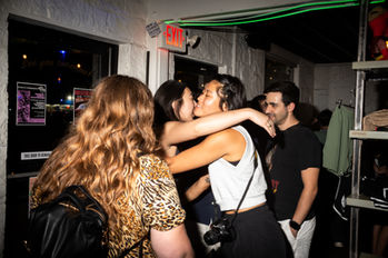 Women embrace and kiss in a crowded club setting, people in background. ATX Event Photography