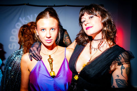 Two women smile at the camera, Spooky Vibes party, wearing stylish dresses.