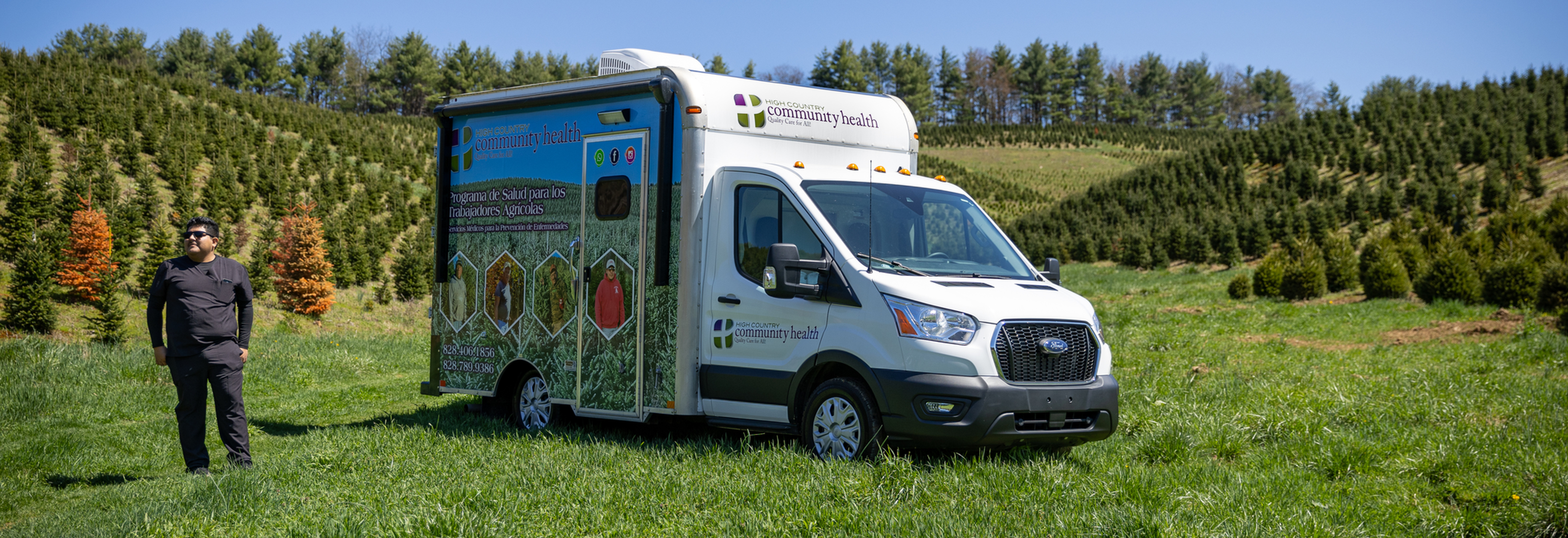 Man stands beside a white High Country Community Health mobile clinic truck in a grassy field with trees under a clear blue sky.