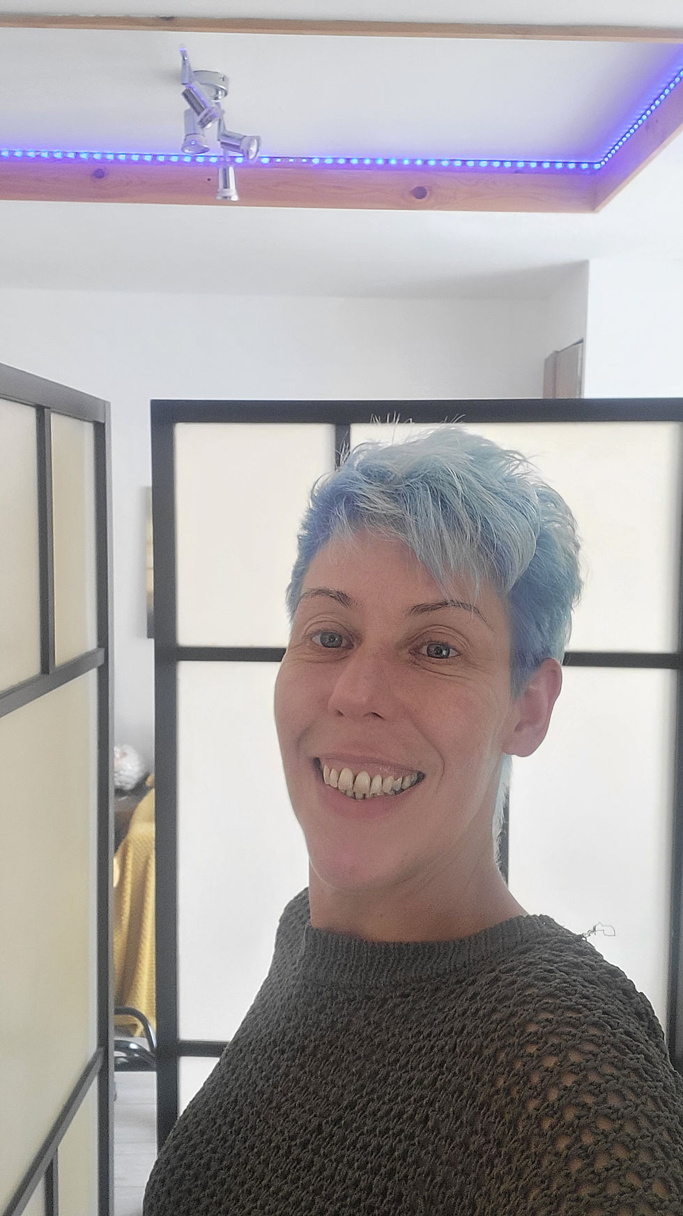 A woman smiles gently while looking at the camera. The background shows a therapy room at Sunshine Holistic Therapy, Manchester