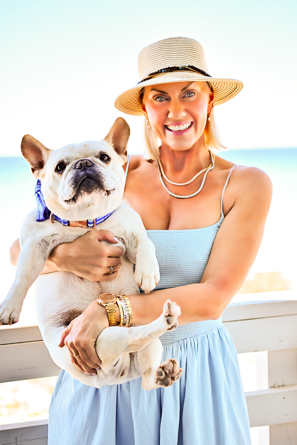 Joanna Olsen smiles while holding her French Bulldog, Utah, on a Gulf Coast balcony overlooking the beach. She wears a light blue sundress, layered jewelry, and a straw hat, reflecting her relaxed coastal lifestyle.