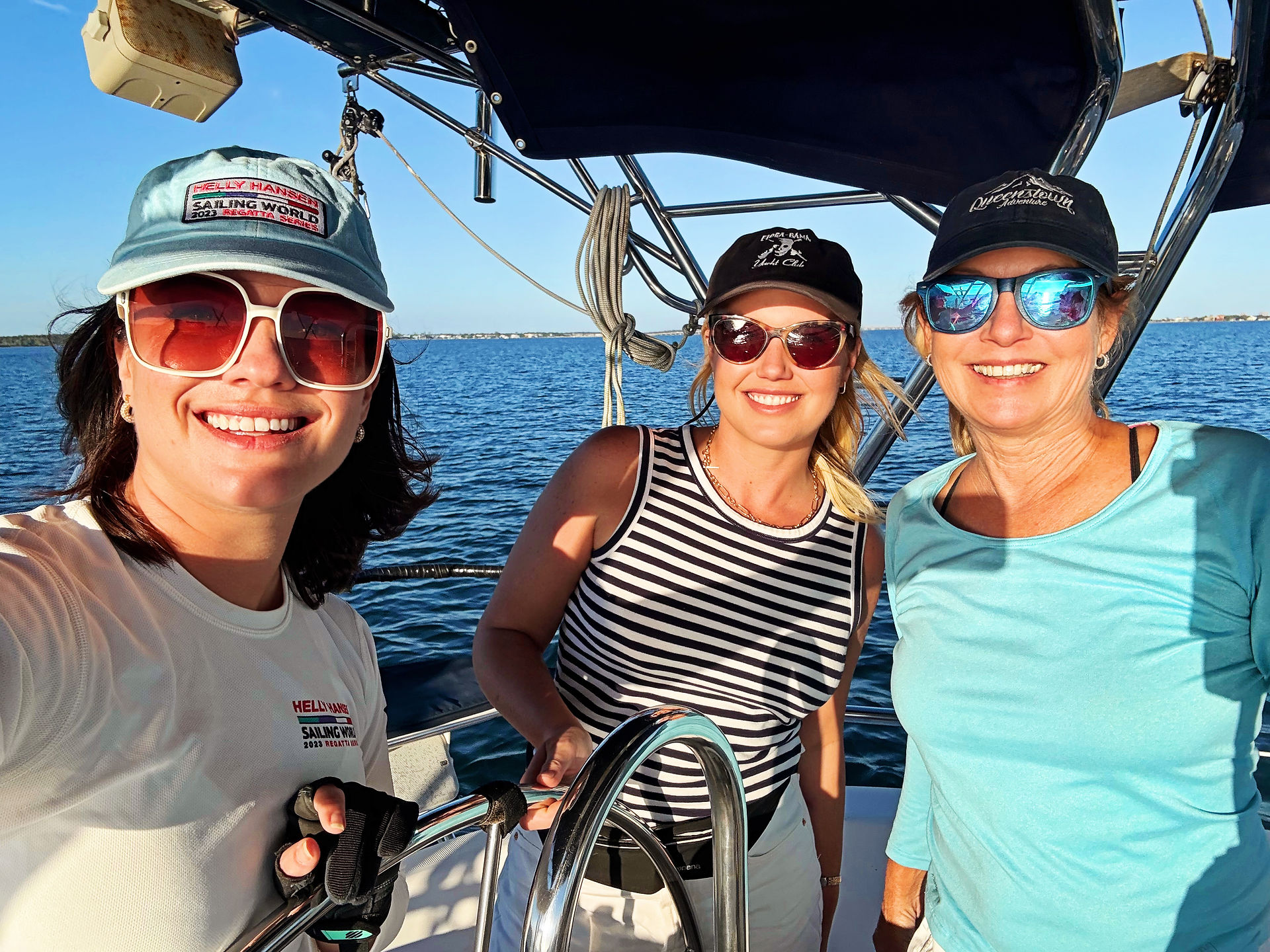 Three women smiling at the helm of a sailboat, wearing sunglasses and casual coastal attire during a day on the water.