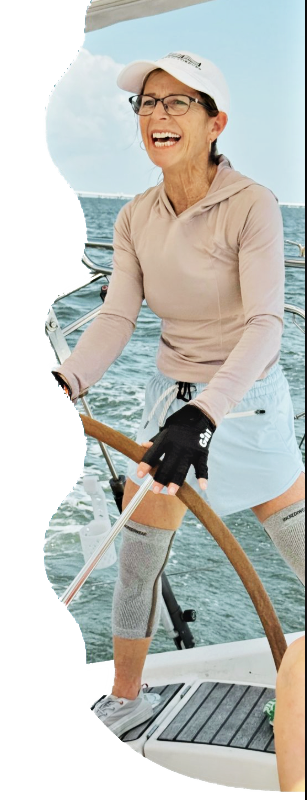 Woman steering a sailboat with focus and confidence, hands on the wheel and open water behind her.