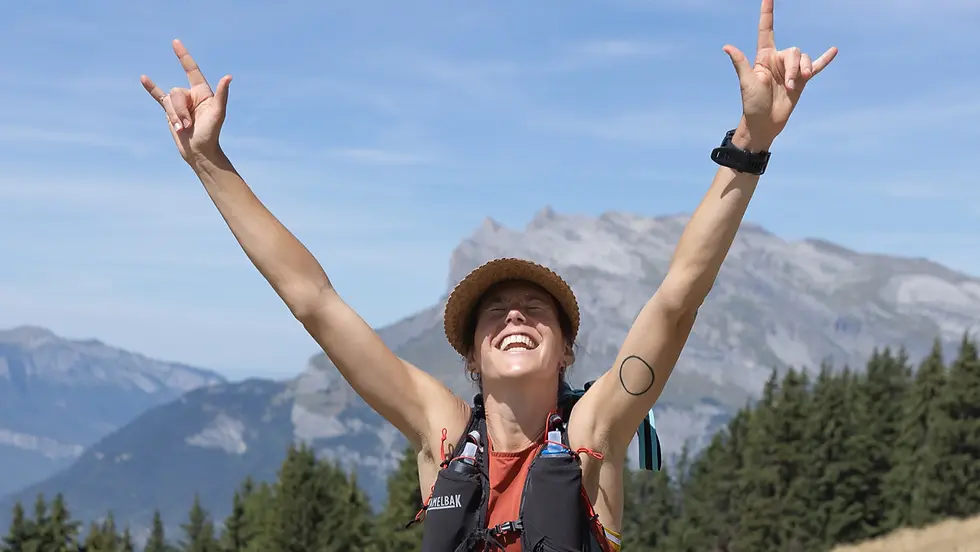 Cat Bradley celebrates on a mountain trail with both arms raised toward the sky, smiling after a successful run. Towering peaks and evergreen forests surround her, reflecting the joy and accomplishment found through endurance sports.