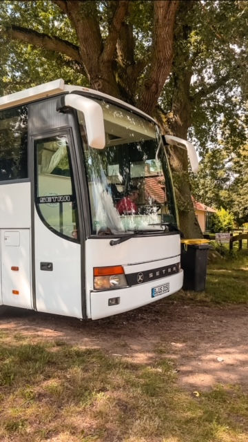 Das Wohnmobil steht bereit zur Abfahrt, wie ein Abenteurer in den Startlöchern. Es parkt mitten im Wald auf einer grünen Rasenfläche, die Umgebung ist ruhig und von hohen Bäumen umgeben. Die Tür des Wohnmobils ist fest geschlossen, als würde es nur darauf warten, sich auf den nächsten Abschnitt der Reise zu begeben. Die Szene strahlt Vorfreude und eine leichte Spannung aus, als stünde das nächste Abenteuer kurz bevor.