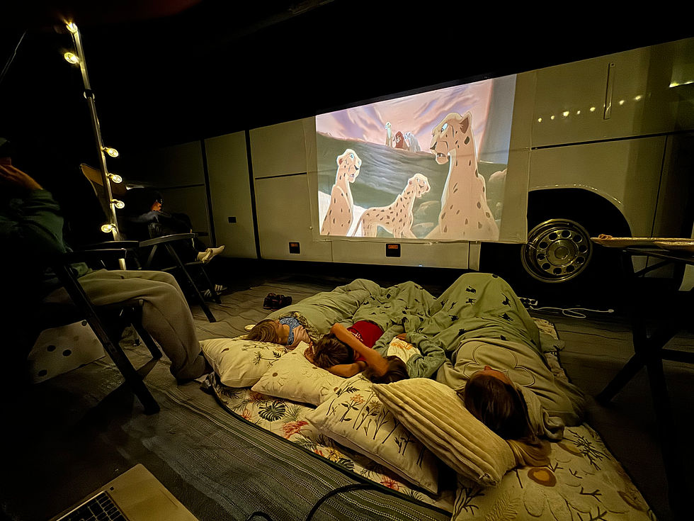 Ein improvisierter Kinoabend auf einem Campingplatz: Eine Filmprojektion mit einer Szene aus einem Zeichentrickfilm wird auf die Seite eines großen weißen Reisebusses projiziert. Vor dem Bus liegen mehrere Kinder und Erwachsene in Decken und Kissen eingekuschelt auf dem Boden, während sie den Film genießen. Links im Bild sind dekorative Lichterketten zu sehen, die eine gemütliche Atmosphäre schaffen. Im Hintergrund sitzen Personen auf Campingstühlen, die den Film ebenfalls verfolgen. Die Szene vermittelt ein Gefühl von Gemeinschaft, Gemütlichkeit und Kreativität, die das Camperleben so besonders machen.