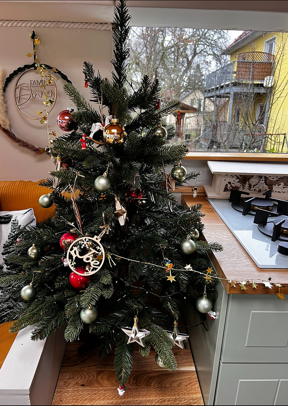Unser Weihnachtsbaum im Donnercamper: Liebevoll geschmückt mit roten, goldenen und silbernen Kugeln sowie filigranen Holzornamenten. Durch das große Fenster fällt Tageslicht auf die gemütliche Wohnküche im selbst ausgebauten Reisebus.