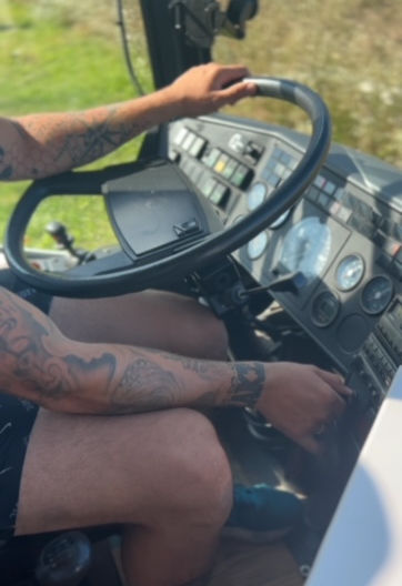 Ein Mann mit tätowierten Armen und kurzer Hose steckt die Autoschlüssel in das Zündschloss, um den Motor seines Campers zu starten. Der Fokus liegt auf dem großen Lenkrad des Busses, während im Hintergrund die Innenausstattung des Fahrzeugs sichtbar ist. Die Szene vermittelt das Gefühl von Abenteuer und Vorfreude auf die bevorstehende Reise.