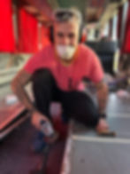 Ein Mann mit Atemschutzmaske, rotem T-Shirt, schwarzen Hosen und blauen Schuhen sitzt auf dem Boden im Inneren eines Campers. In seiner rechten Hand hält er eine Flex. Um ihn herum sind die Fenster des Campers, die mit roten Gardinen zugezogen sind. Auf seinem Kopf trägt er eine Schutzbrille, was auf die handwerklichen Arbeiten hinweist, die er ausführt. Die Atmosphäre vermittelt den Eindruck eines aktiven Ausbaus oder Renovierungsprojekts im Fahrzeug.