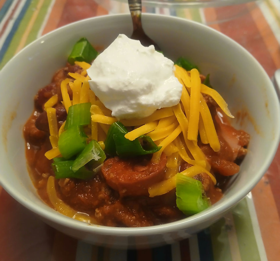 Bowl of chili topped with cheddar, green onions, and sour cream on a striped tablecloth. Warm, appetizing presentation.