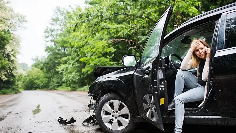 Frustrated woman sitting in damaged car after hit-and-run accident – vehicle front end crushed, driver holding head in distress.