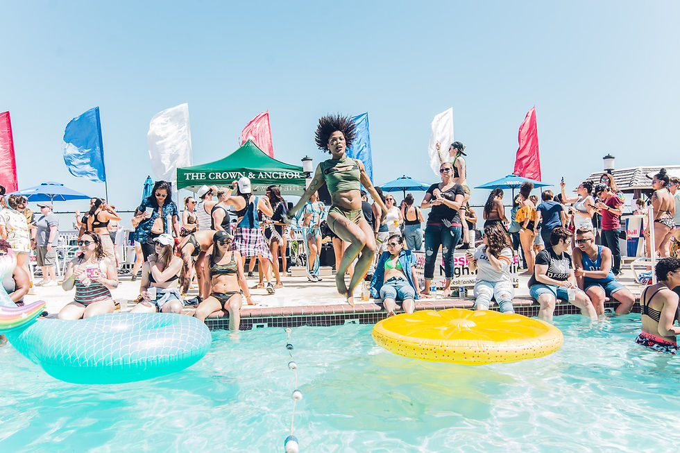 People enjoying a pool party at Memorial Day PTown celebration. A woman jumps into the water, surrounded by colorful floats and flags. A green canopy reads "The Crown & Anchor." Mood: lively.