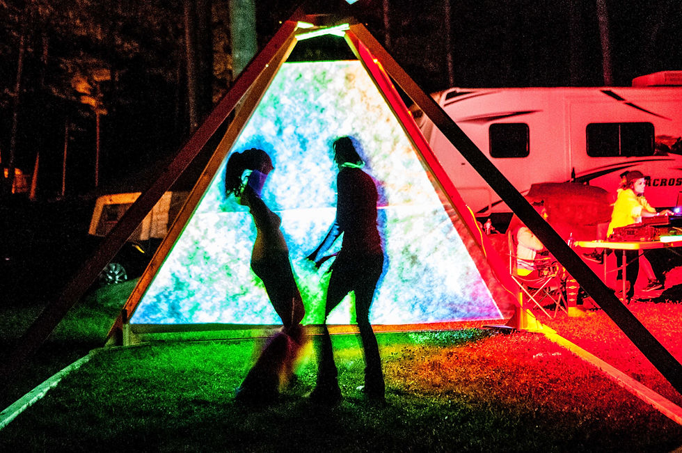 Two silhouettes dance in front of a colorful, lit pyramid structure at night. An RV and people sitting at a table are in the background.