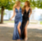 Zwei Frauen in eleganten Abendkleidern, eine in blau, die andere in schwarz, gehen Arm in Arm im Freien unter Bäumen