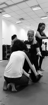 Foto 5: uma mulher, vista de costas, está sentada sobre os calcanhares e mexe na perna de uma outra mulher que está diante dela, ajoelhada. Ao fundo, há outras pessoas em pé.