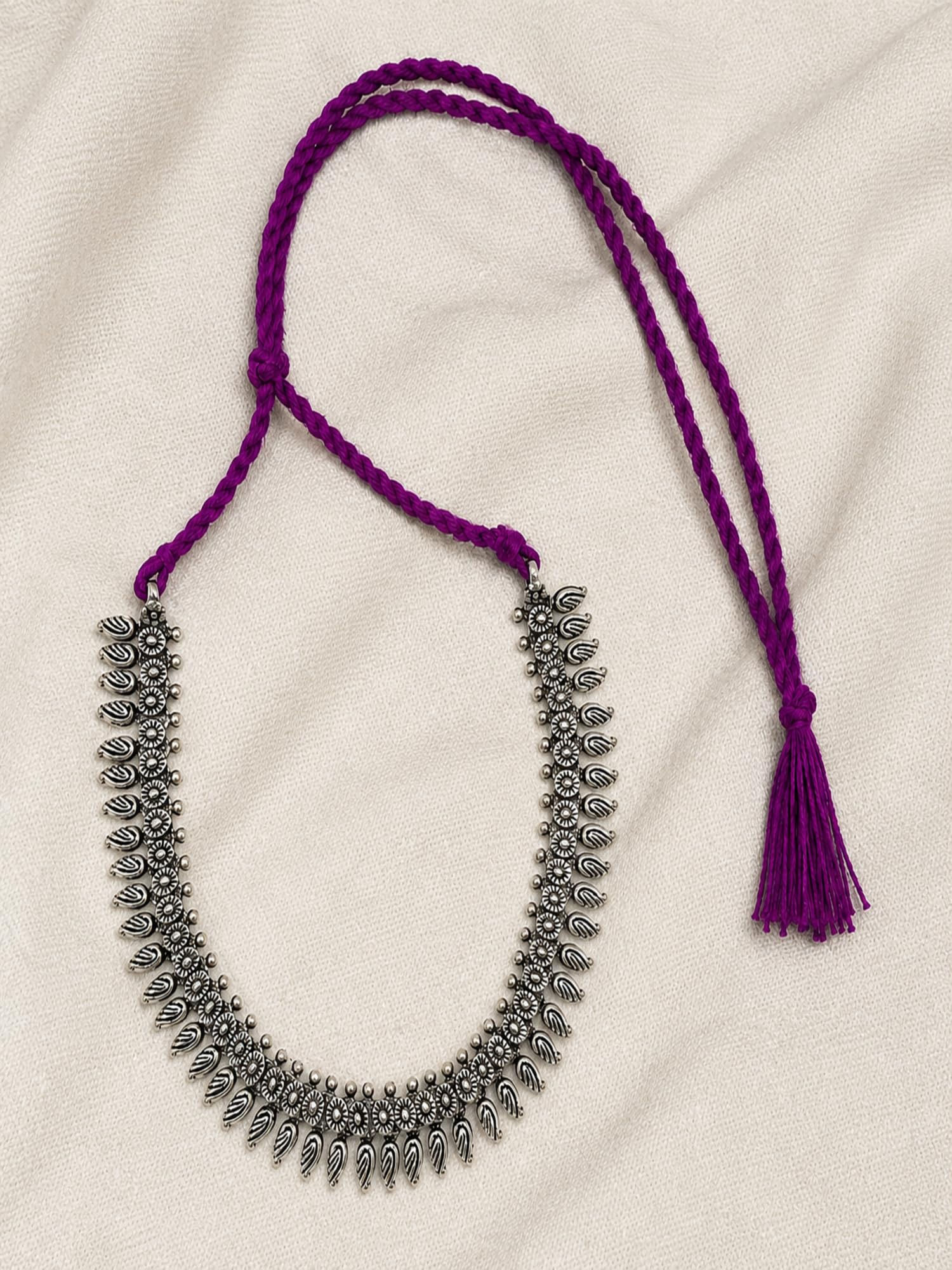 Athizay oxidised silver necklace featuring floral motifs, dangling charms, purple braided thread and tassel on a fabric backg