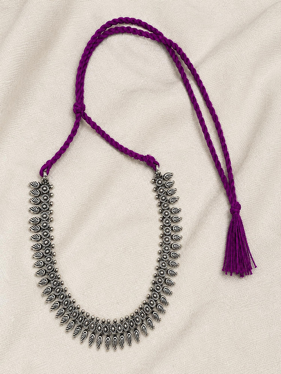 Athizay oxidised silver necklace featuring floral motifs, dangling charms, purple braided thread and tassel on a fabric backg