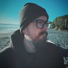 Bearded man in glasses and a beanie looks thoughtful on a pebbled beach with blue skies and green hills in the background.
