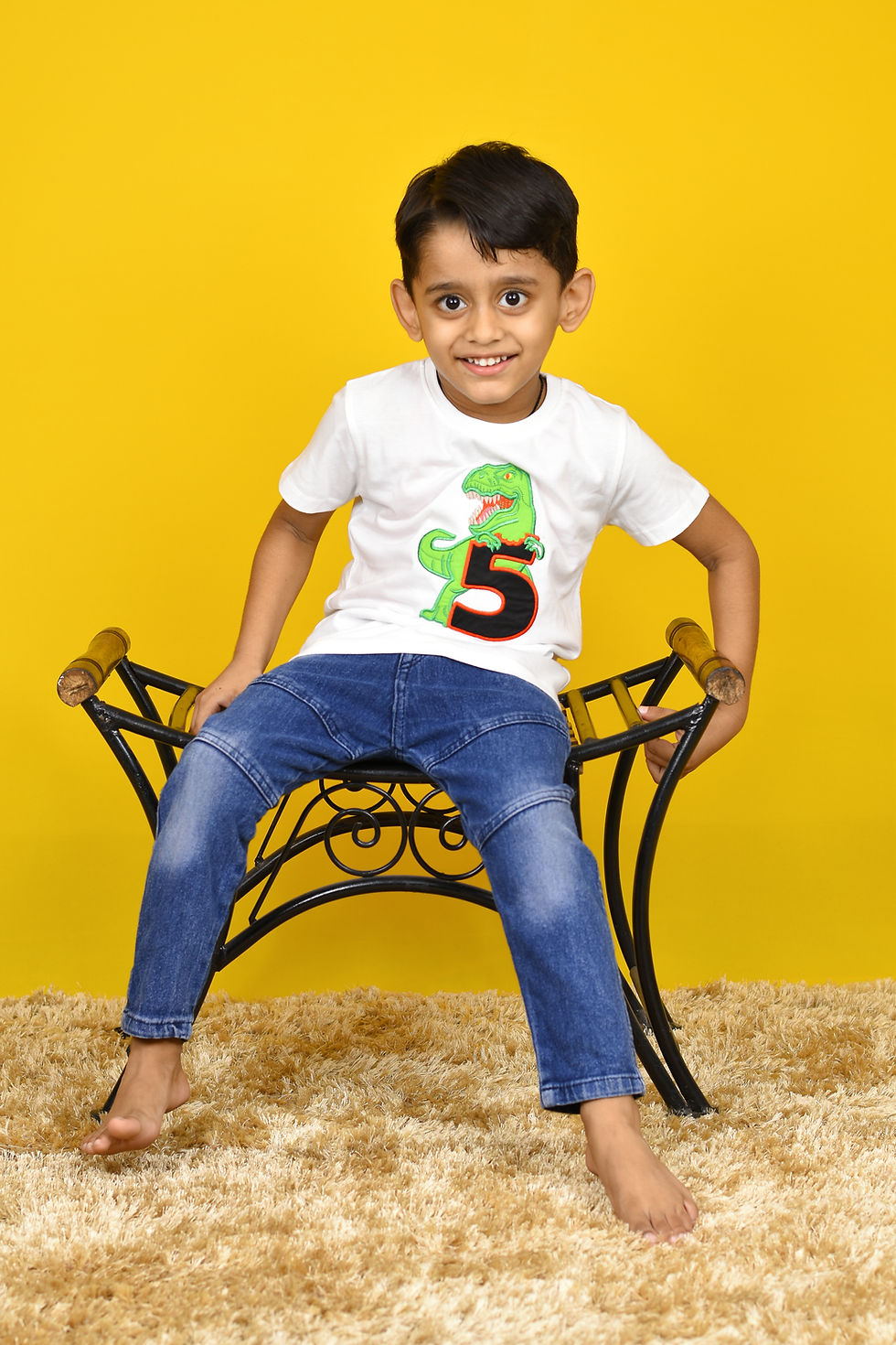 Thumbnail: another photo of a kid sitting and wearing a t shirt by Hugs&Hues having a dinosaur and number 5 embroidered on it and posing